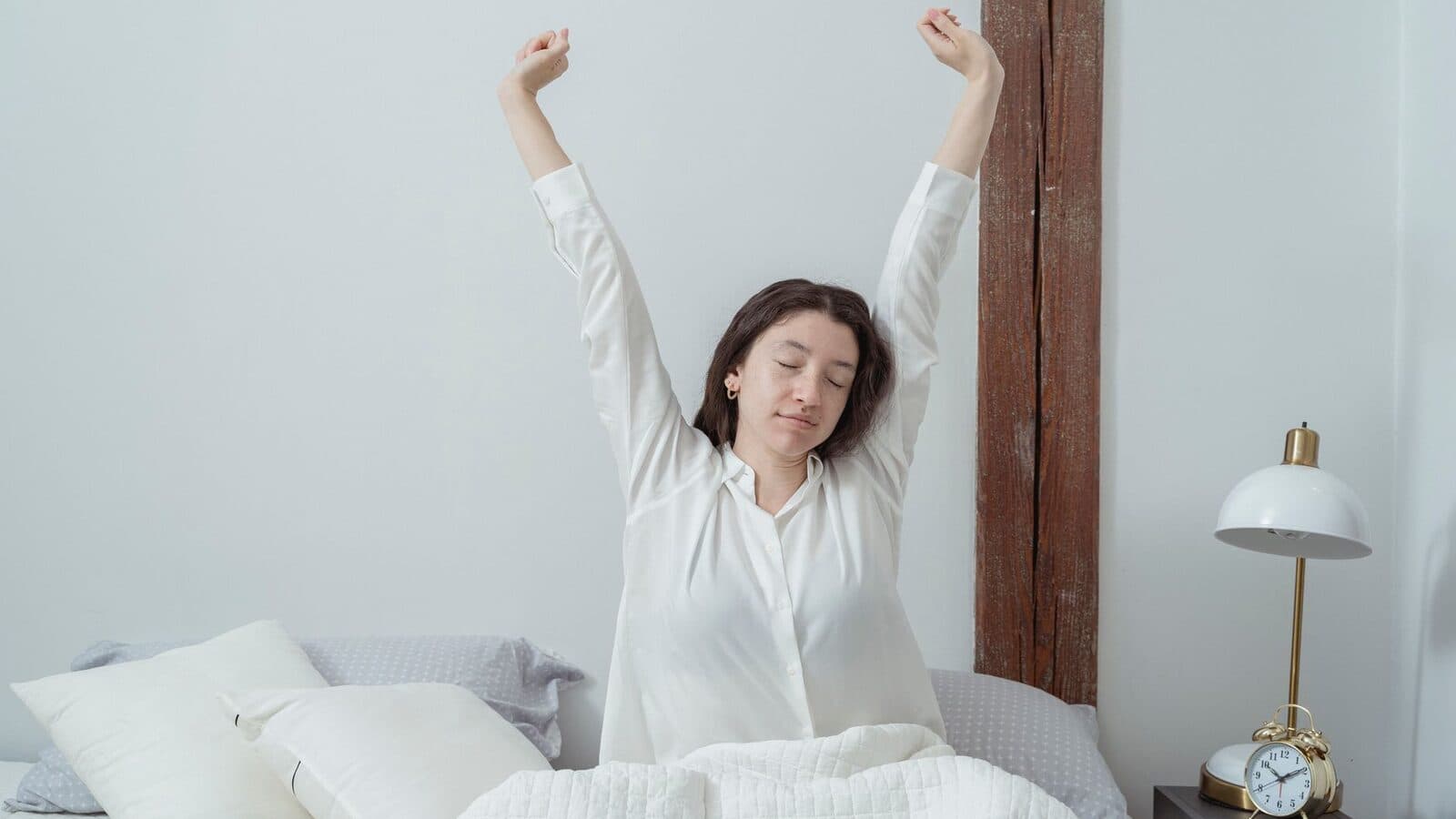 Vrouw rekt zich uit in bed naast een lamp en een wekker